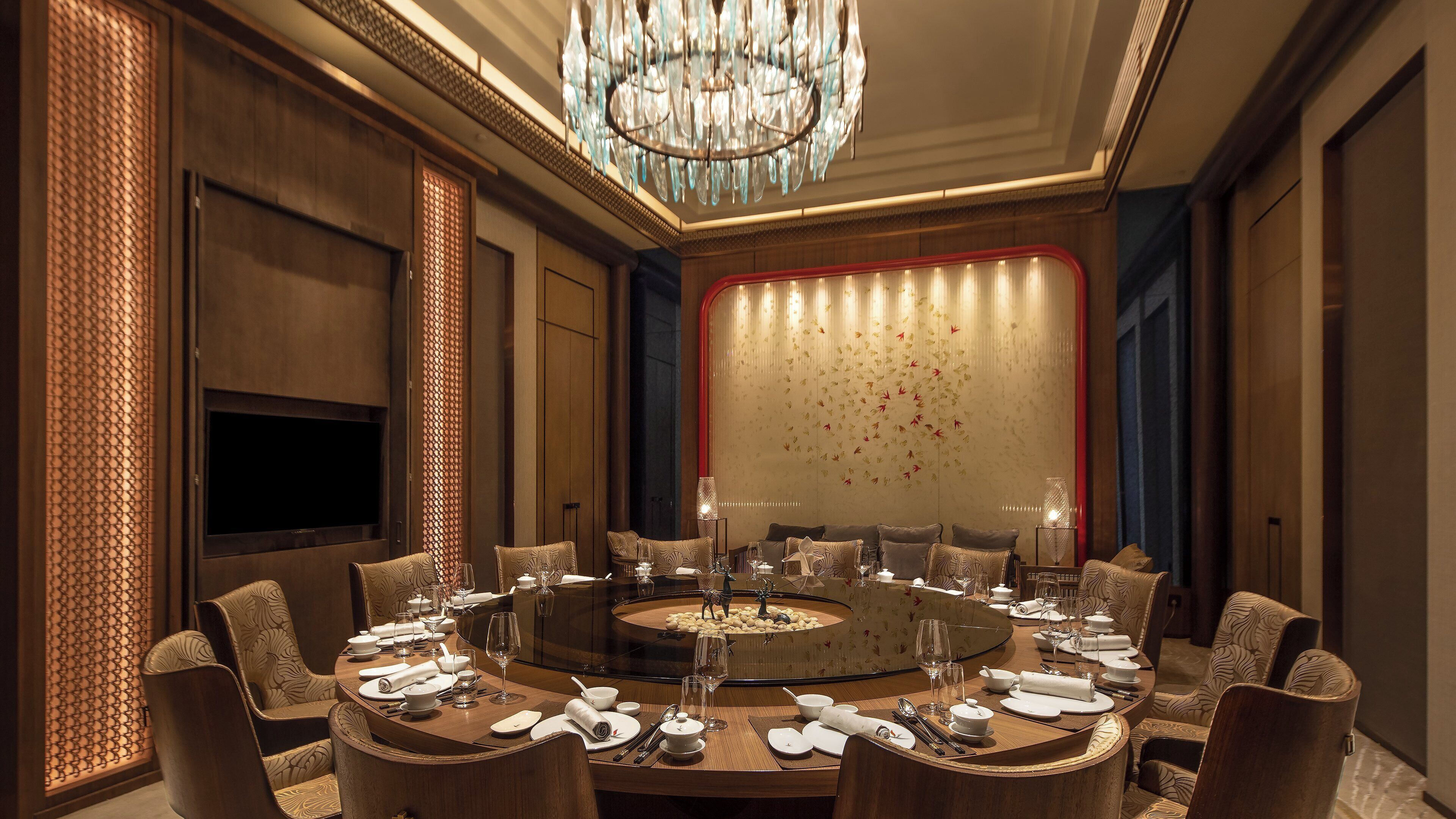 A round dining table with chairs, a chandelier, and a decorative wall with leaf patterns.