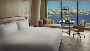 A hotel room with a large bed, a sofa, a desk with a vase of flowers, and a view of a city skyline and water. - InterContinental Tianjin Yujiapu Hotel & Residences by IHG (Tianjin)