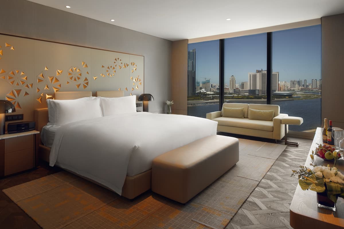 A modern hotel room with a large bed, a sofa, a desk with a vase of flowers, and a view of a city skyline.
