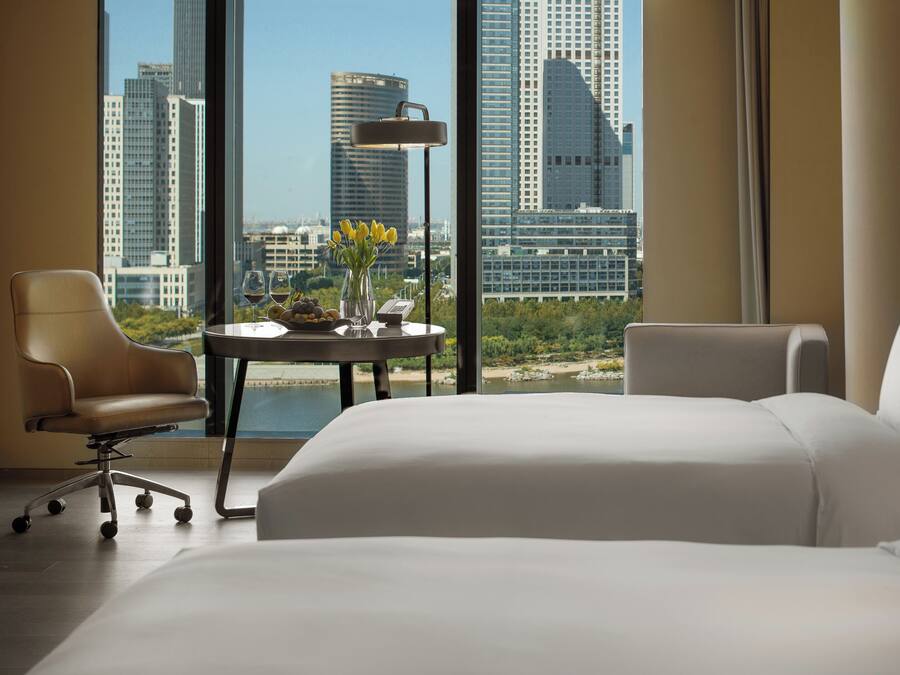 A hotel room with a bed, two chairs, a table with a vase and fruit, a lamp, and a view of city buildings.