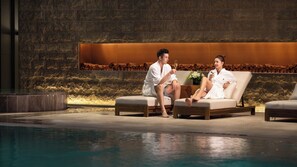 Two individuals seated on a lounge chair by a poolside, with a stone wall and logs in the background.