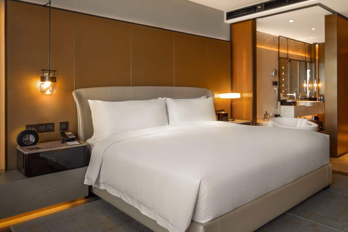 A modern hotel room with a large bed, bedside tables, a desk, and a bathroom with a bathtub and mirror.