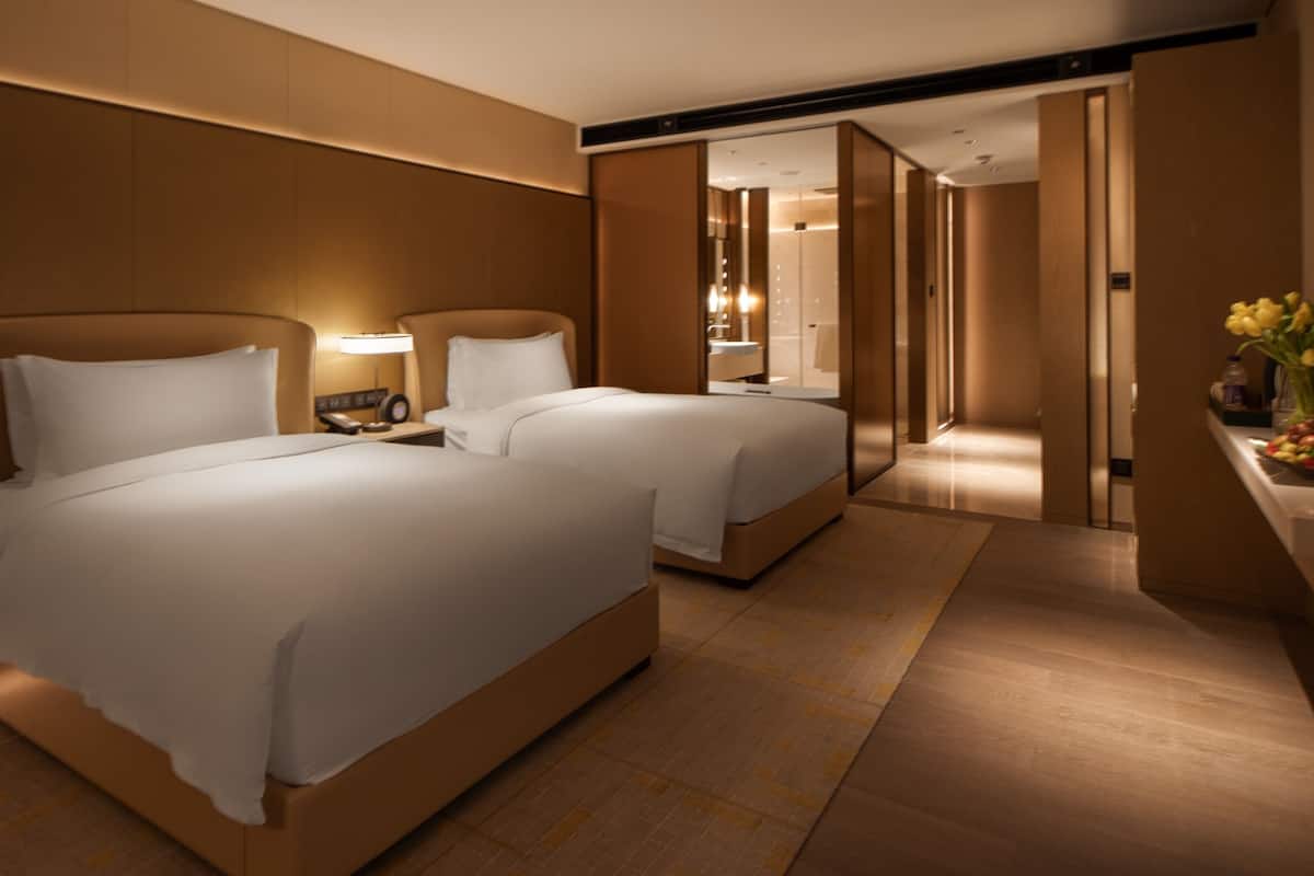 A hotel room with two beds, a bedside table with a lamp, a small table with fruits and a vase of flowers, and a view into the bathroom.