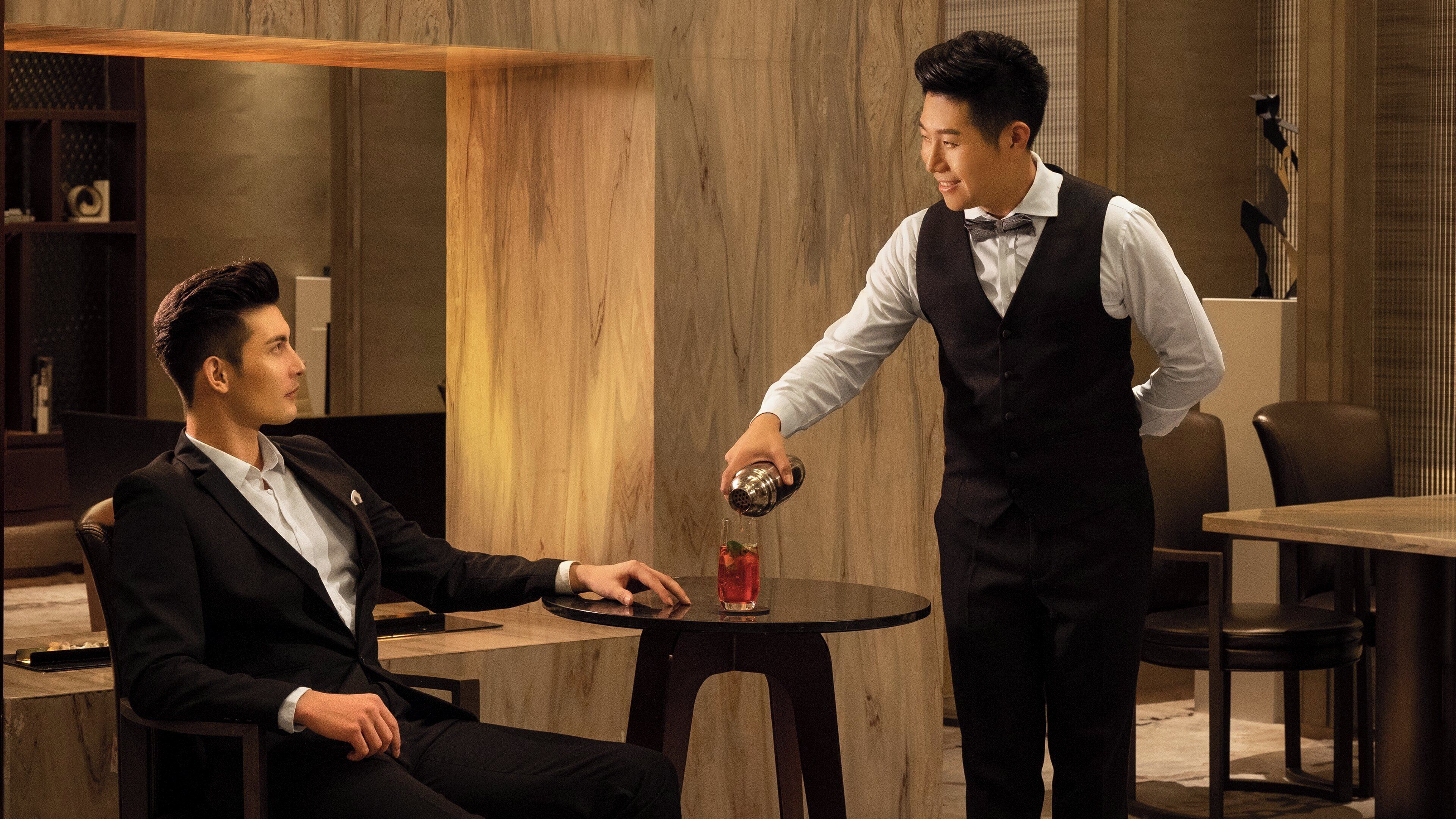 A man in a suit sitting at a table with a drink, another man in a vest pouring a drink into it.