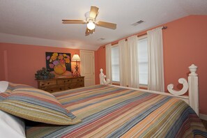 Une chambre à coucher comprenant un lit, un ventilateur de plafond, une commode, un miroir et une fenêtre avec des rideaux.