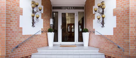 A hotel entrance with a sign reading "HOTEL" above the door, flanked by two large potted plants and wall-mounted light fixtures.