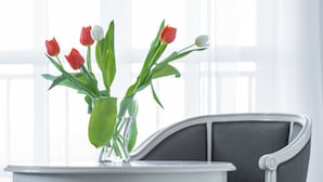 Uma mesa redonda branca com um vaso de tulipas, uma cadeira estofada cinzenta e uma janela com cortinas leves ao fundo.
