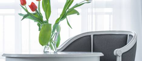 A white round table with a vase of tulips, a grey upholstered chair, and a window with sheer curtains in the background.