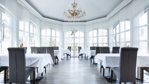 Uma ampla sala de jantar com mesas brancas e cadeiras cinzentas, grandes janelas e um lustre.