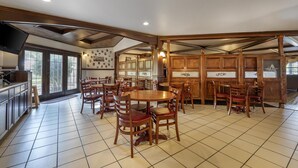 Un vaste espace de salle à manger avec des tables et des chaises en bois, un coin cuisine équipé d’un téléviseur à écran plat, et de grandes fenêtres laissant entrer la lumière naturelle.
