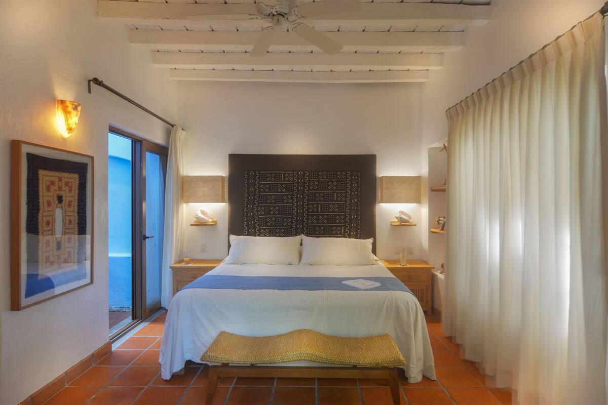 Un dormitorio con cama, cabecera, lámparas de noche, ventana y puerta.