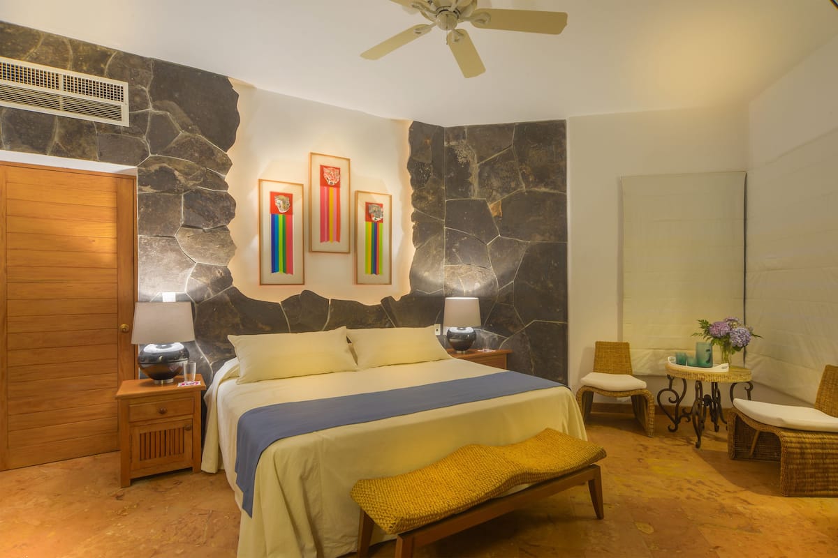 Un dormitorio con cama, mesitas de noche, una puerta de madera, un muro decorativo de piedra, un ventilador de techo y una pequeña mesa con un florero.