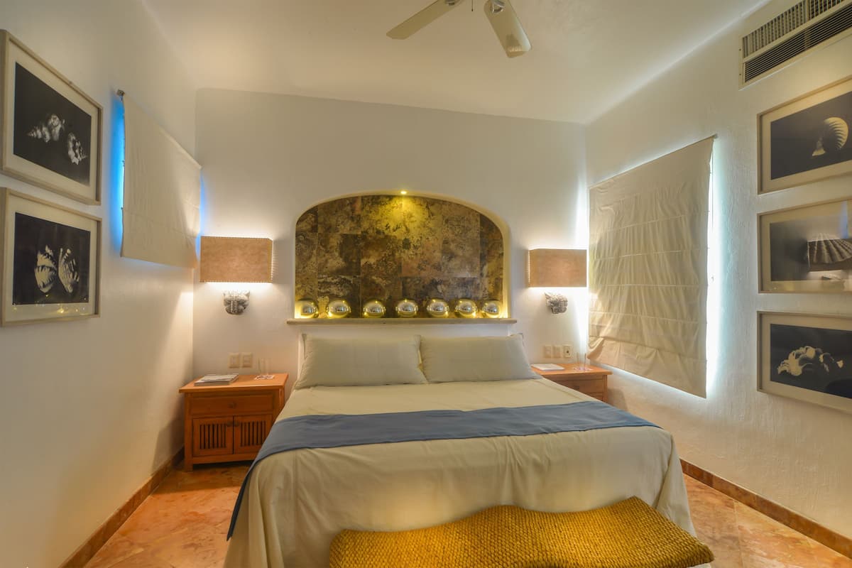 Una habitación de hotel con una cama grande, dos mesitas de noche, un ventilador de techo y cuadros en las paredes.