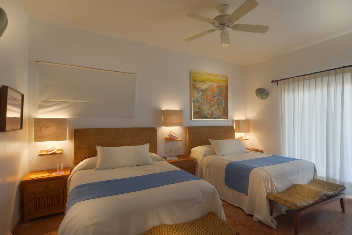 Habitación de hotel con dos camas, un ventilador de techo, un cuadro y una ventana con cortinas.