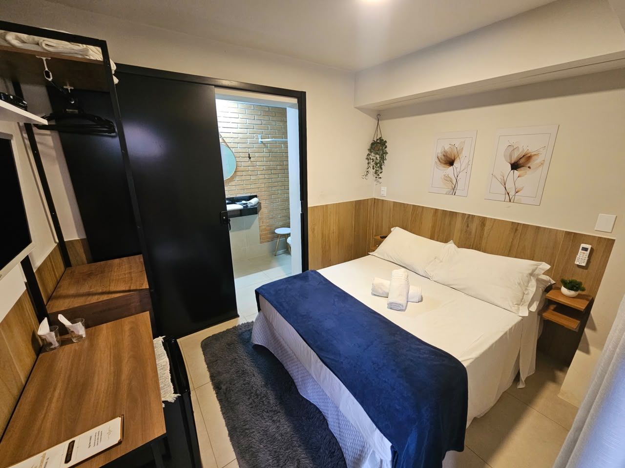 Une chambre d’hôtel avec un lit, une table de chevet et un petit bureau avec une chaise. Une porte donne accès à une salle de bain et à un espace douche.