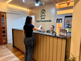 Uma receção com o letreiro 'Hotel Casa Encantada', um balcão com visor digital, um frigorífico abastecido com bebidas e uma câmara de segurança no alto.