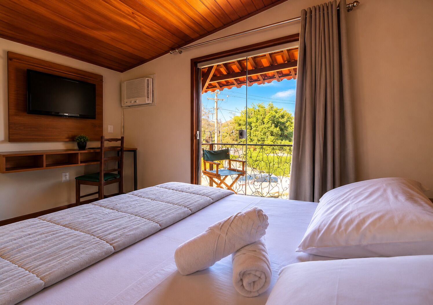 Une chambre d’hôtel avec un lit, un téléviseur fixé au mur, un balcon avec une chaise et une fenêtre avec des rideaux.