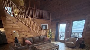 A cozy wooden interior with a staircase, leather sofas, a coffee table, and a small Christmas tree. - Deep Creek Overlook Cabin HGTV Log Cabin Living WiFi Pool Table Hot Tub King Bed (Bryson City)