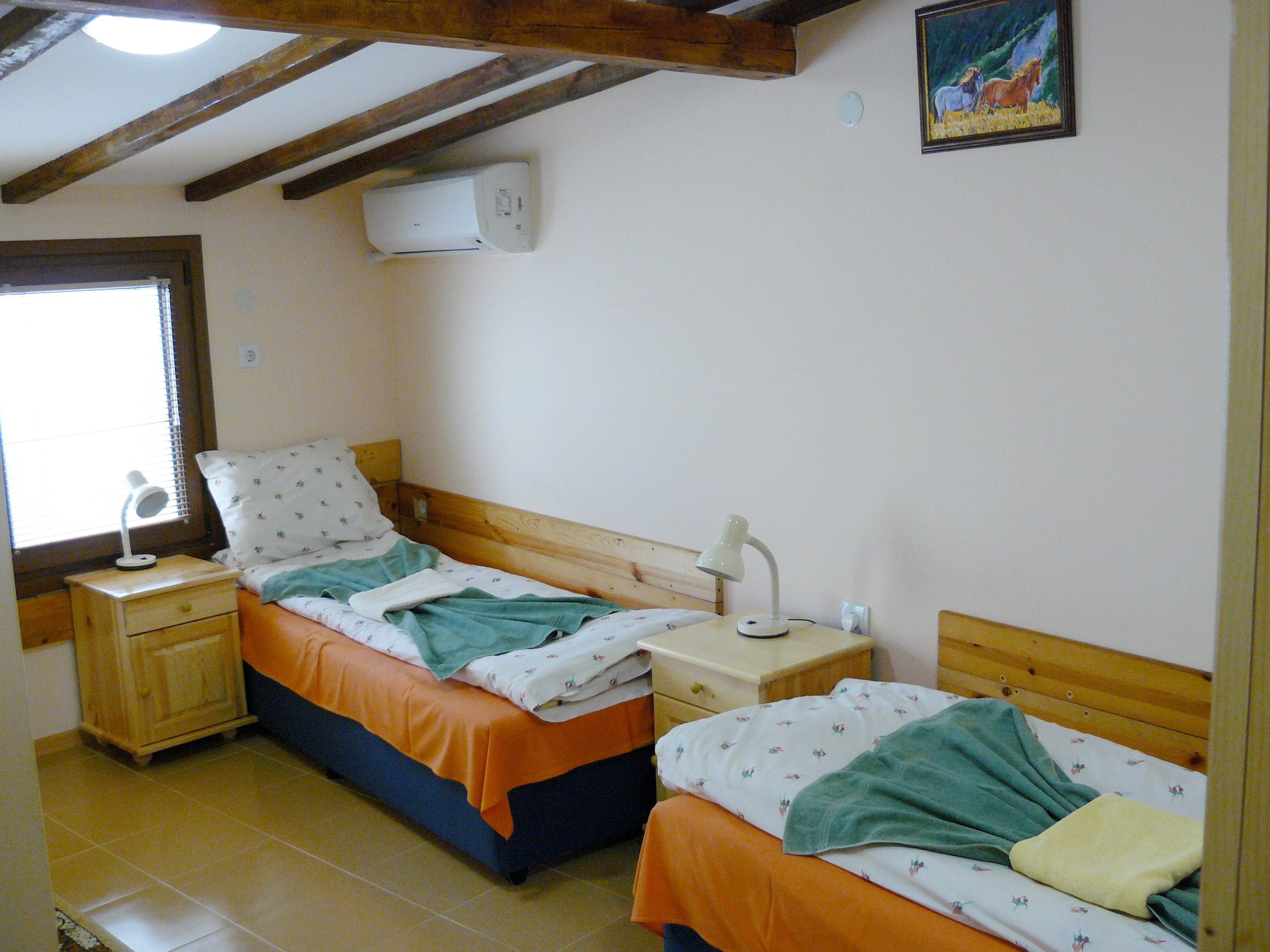 A small, cozy bedroom with two single beds, wooden headboards, a window with blinds, a wall-mounted air conditioner, and a framed picture on the wall.
