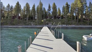 Un muelle de madera que se adentra en un cuerpo de agua, rodeado de un bosque denso y un edificio al fondo.