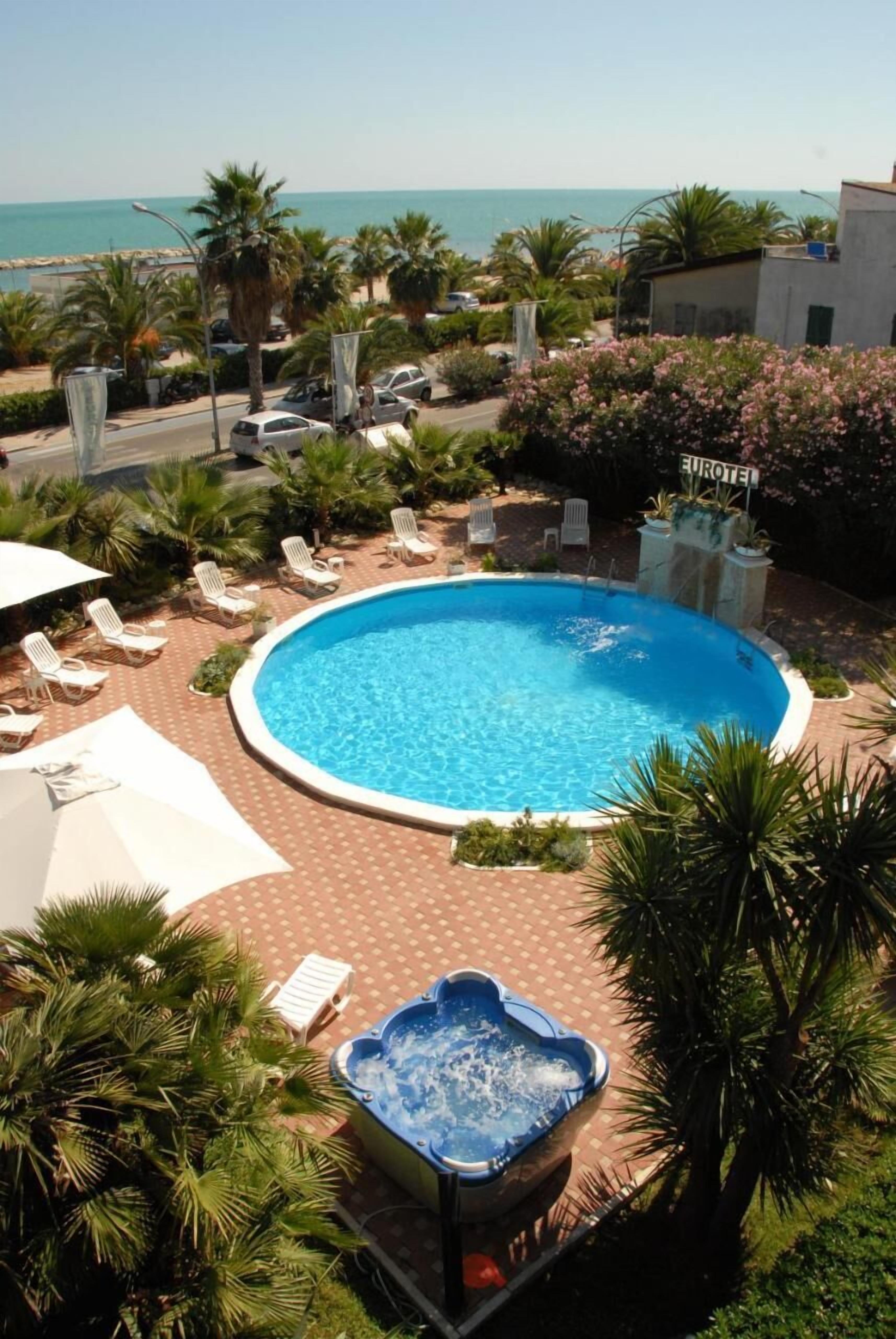 A circular pool with lounge chairs and a hot tub, surrounded by palm trees and a view of the sea.