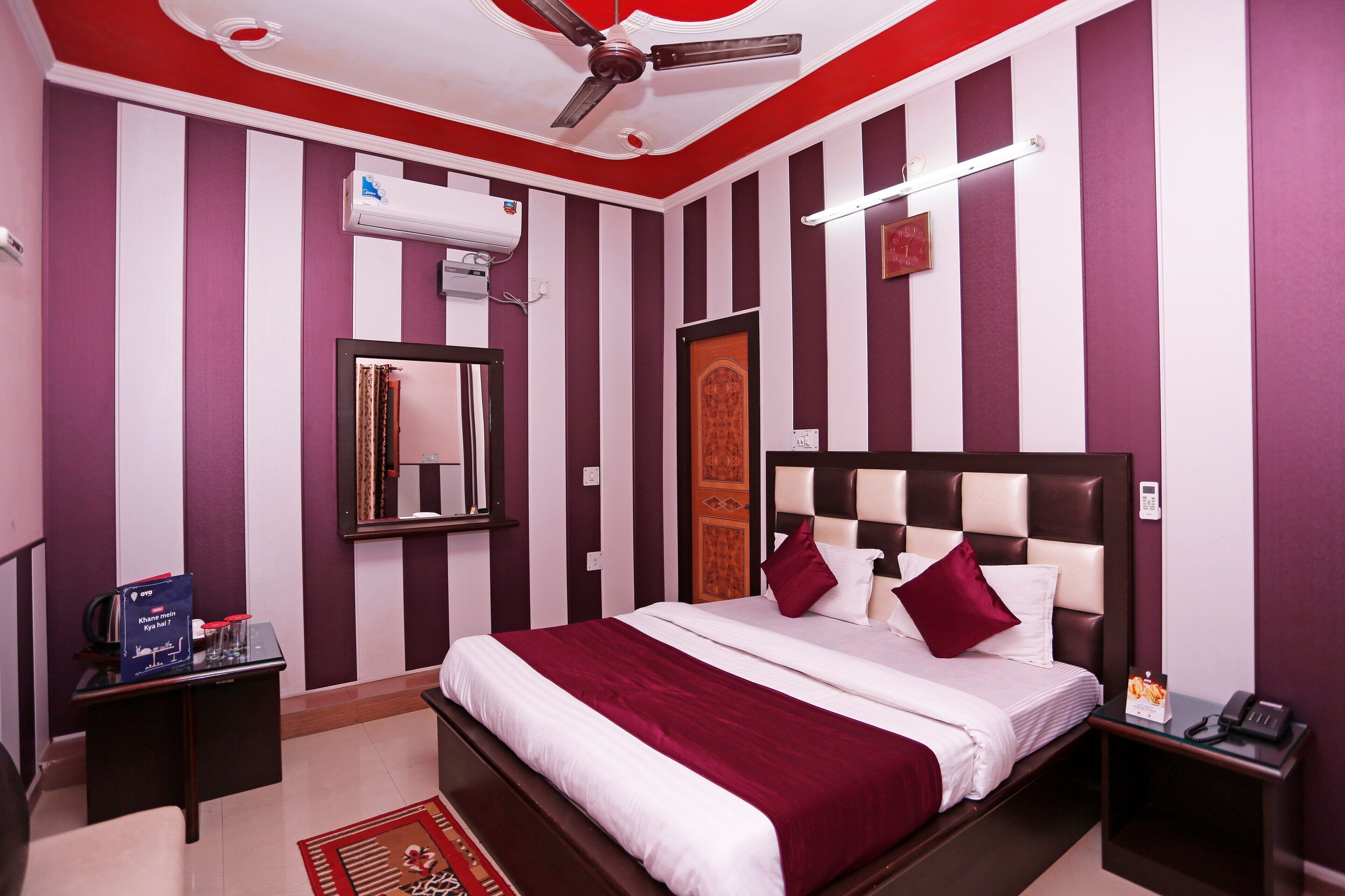 A hotel room with a striped wall design, a bed with white and maroon bedding, a ceiling fan, an air conditioning unit, a bedside table with a phone, and a small table with a book and a card.
