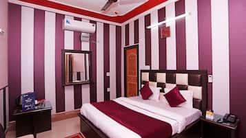 A hotel room with a striped wall design, a bed with white and maroon bedding, a ceiling fan, an air conditioning unit, a bedside table with a phone, and a small table with a book and a card.