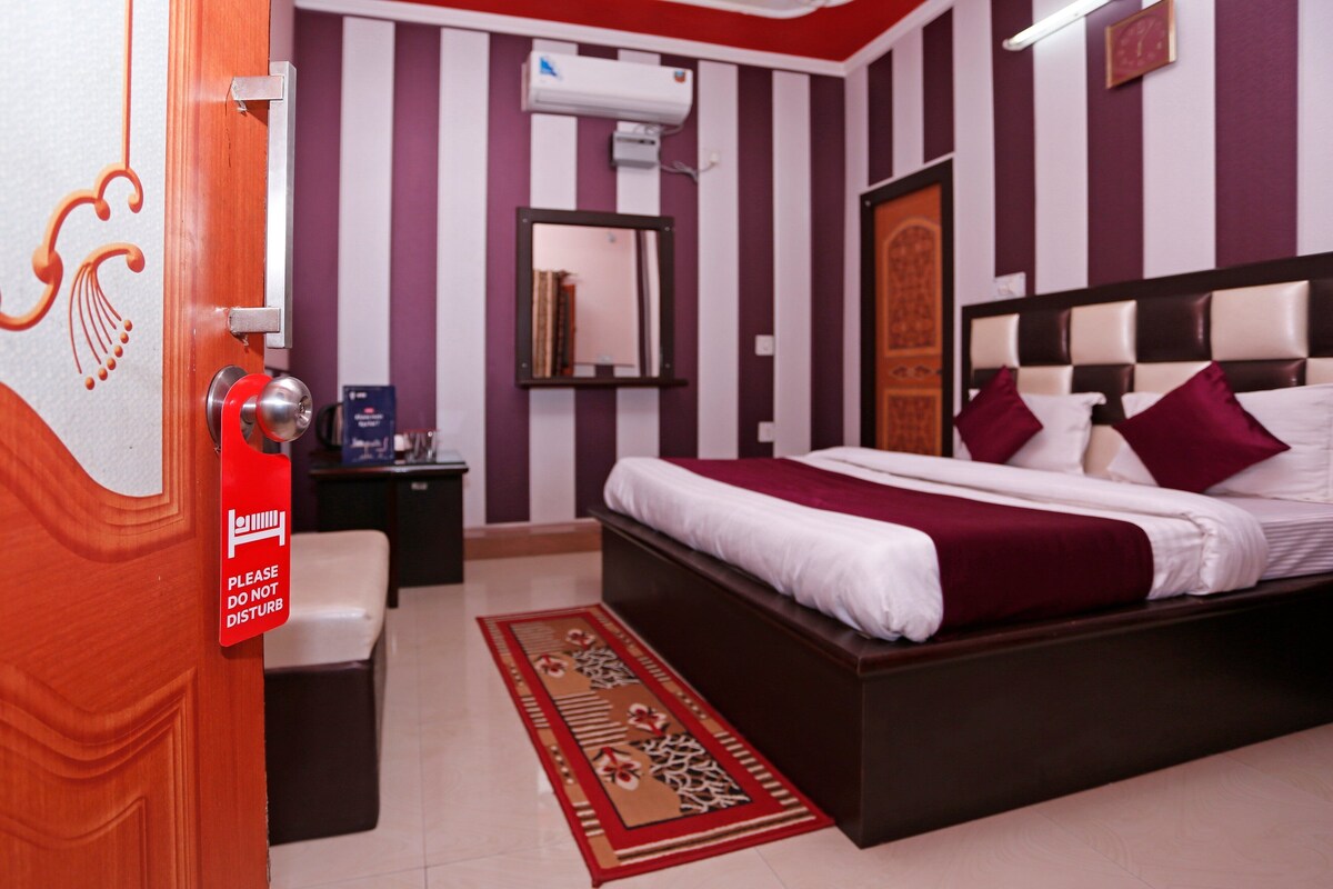 A hotel room with a bed, a nightstand, a mirror, and a red sign on the door.