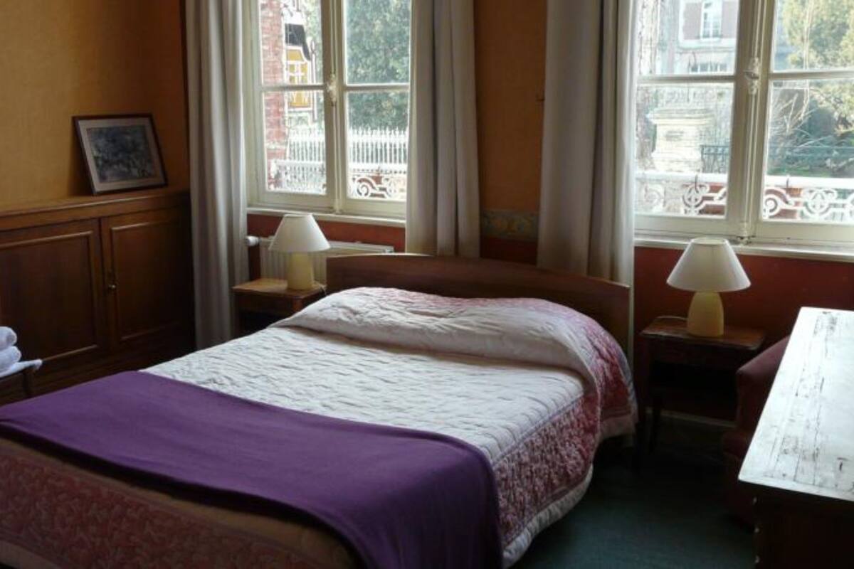 Un dormitorio con cama, dos mesitas de noche con lámparas, una silla, armario y ventana con vistas a edificios.