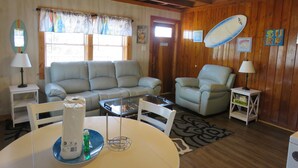 A cozy living room with a sofa, armchairs, a coffee table, and a dining table. There are surf-themed decorations and a ceiling fan. - Summer Special/RIGHT ACROSS from OCEAN/Fenced yard/pet friendly  (Nags Head)
