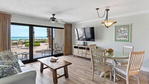Sala de estar con televisor, mesa de comedor y sillas, un sofá, y vista a la playa a través de amplios ventanales.