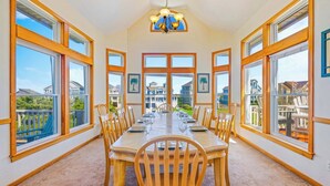 A dining room with a long table, multiple chairs, and large windows offering views of the neighborhood. - Semi-Oceanfront Dog Friendly w/ Pool, Hot Tub, Home Theater, and Game Room (Avon)