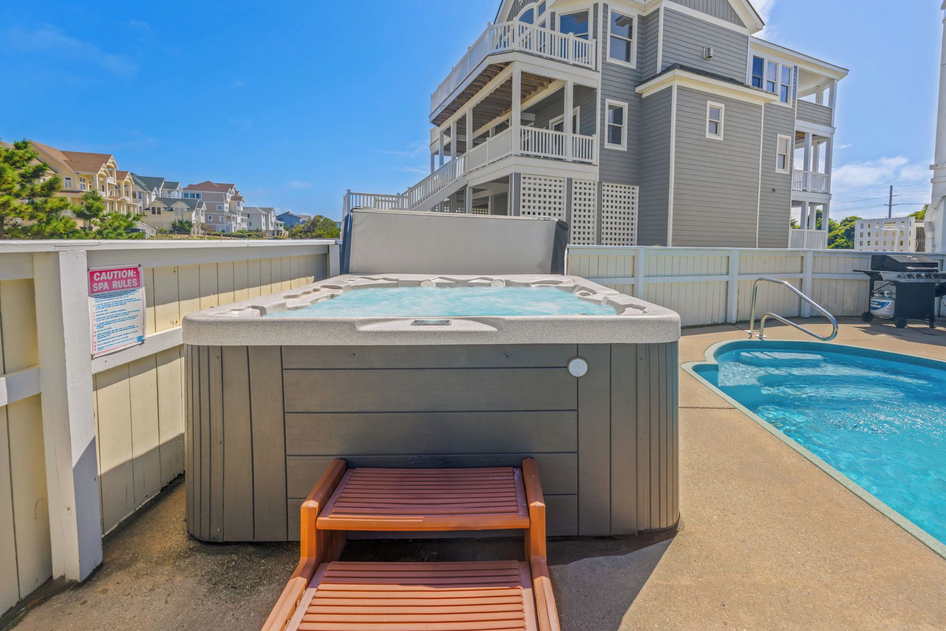 A hot tub with a wooden bench in front, a pool, and a multi-story building in the background.