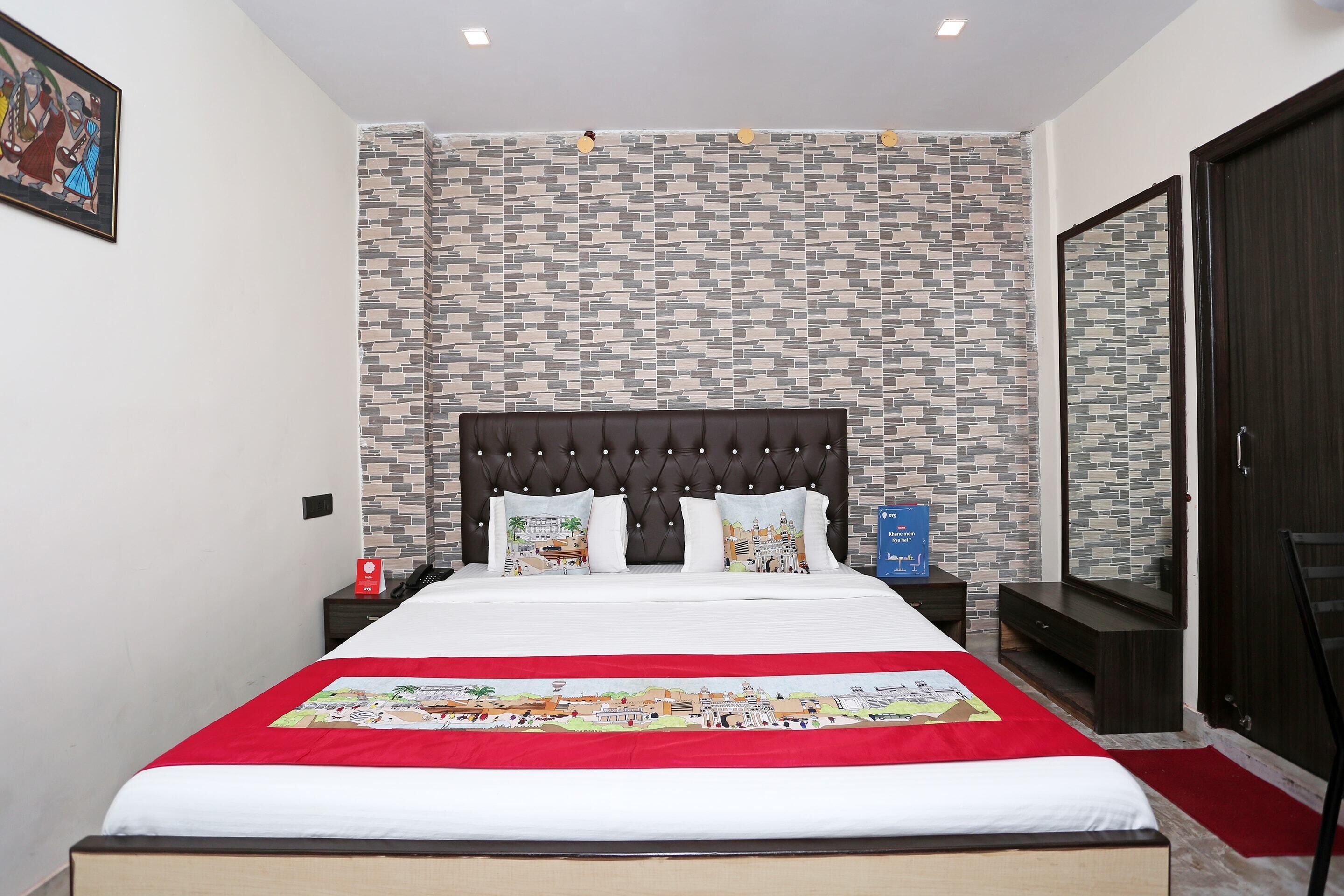 A neatly arranged hotel room with a large bed, a headboard, a nightstand, a dresser, a mirror, and a red carpet.