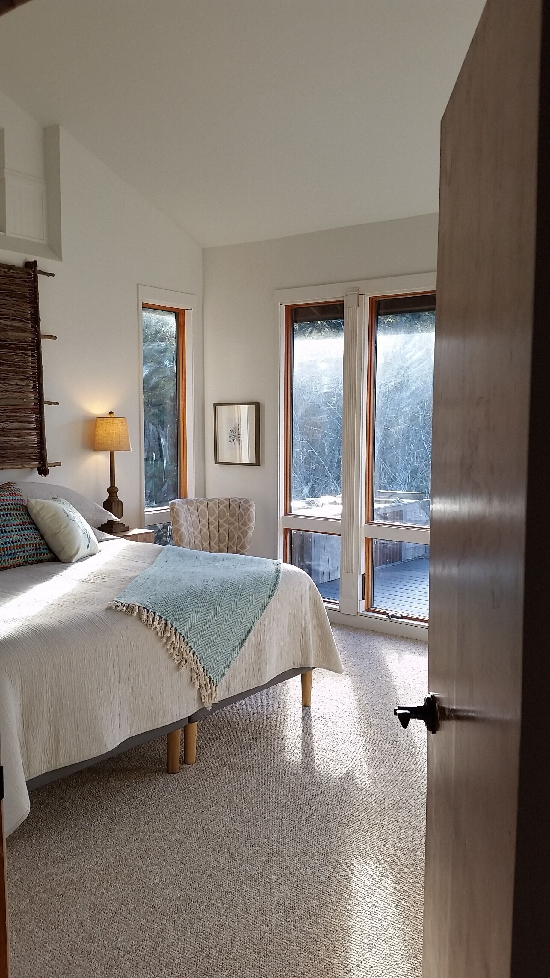 A bedroom with a bed, a nightstand, a lamp, a chair, and a large window with a view of trees.