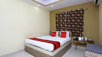 A hotel room with a bed, a bedside table, a small table with a basket, and an air conditioning unit.