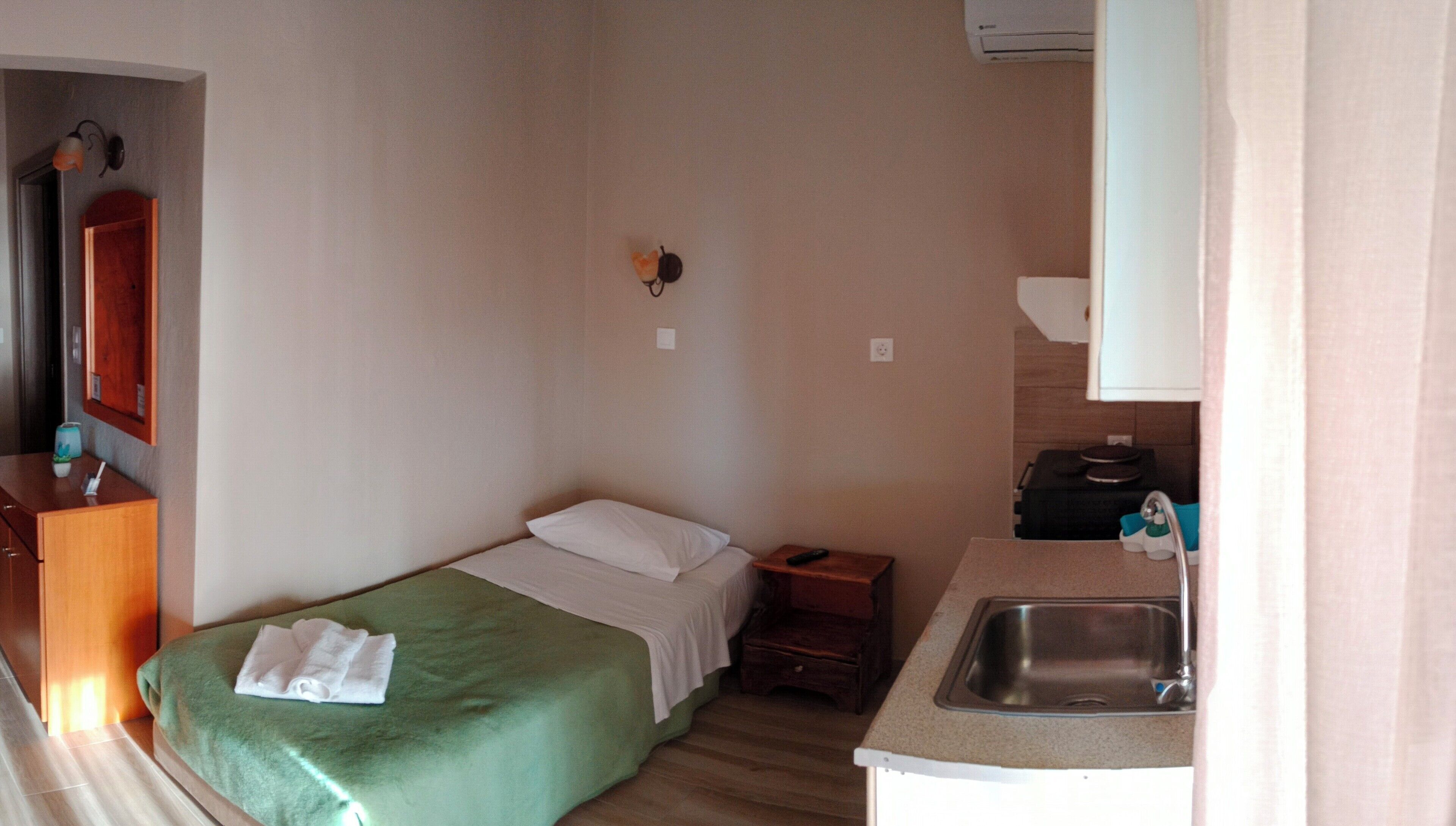 A small, single-bed room with a green bedspread, a wooden nightstand, a small table with a remote control, a sink, and a mirror above the mirror.