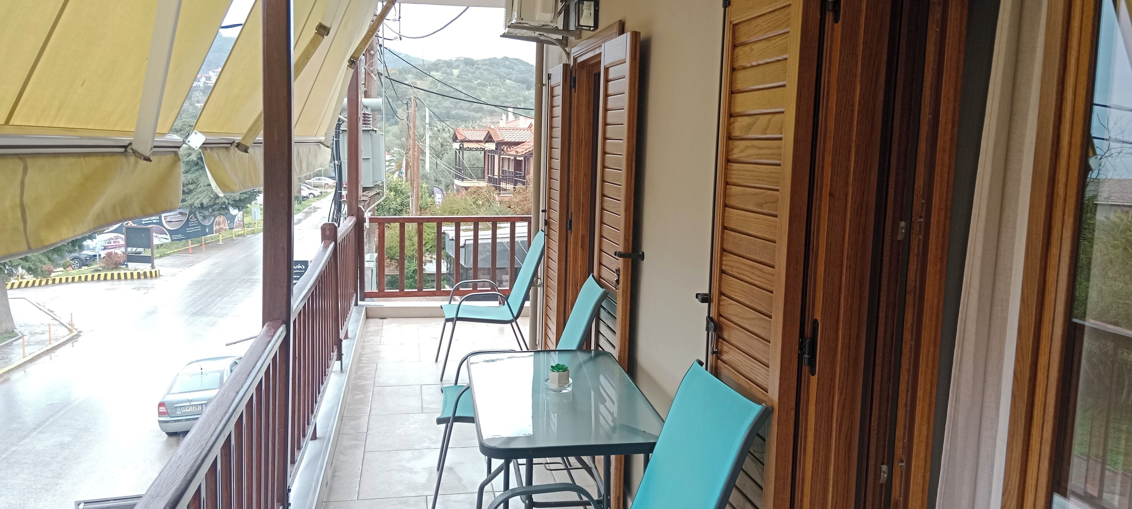 A balcony with a table and chairs, wooden shutters, and a view of the street and buildings.