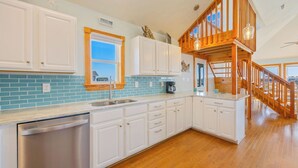 A kitchen with white cabinets, a stainless steel dishwasher, a blue-tiled backsplash, and a wooden staircase. - Oceanview Dog Friendly Home with Private Pool, Hot Tub, Elevator, and Game Room (Waves)