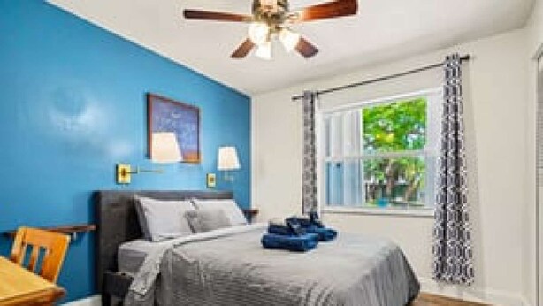 Une chambre à coucher avec un lit, un ventilateur de plafond, une fenêtre avec des rideaux et une vue sur la verdure à l’extérieur.