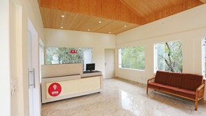 A spacious lobby with a wooden ceiling, a reception desk, a sofa, and large windows offering views of trees. - OYO 9485 Swiss Camps (Kullu)