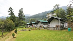 A series of rustic, single-story cabins with green roofs, set in a lush, green valley with mountains in the background. - OYO 9485 Swiss Camps (Kullu)