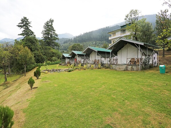 A series of rustic, single-story cabins with green roofs, set in a lush, green valley with mountains in the background. - OYO 9485 Swiss Camps (Kullu)