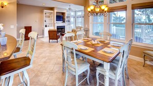 A dining area with a wooden table set for six, surrounded by white chairs with cushioned seats. - Oceanview Home with Hot Tub, Game Room, Wet Bar, Grill. Feel That Summer Breeze! (Salvo)