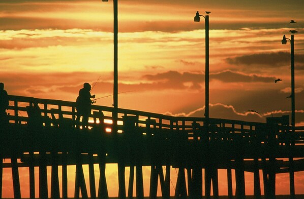 A silhouette of a person fishing on a pier with a sunset in the background. - Oceanview Home with Hot Tub, Game Room, Wet Bar, Grill. Feel That Summer Breeze! (Salvo)