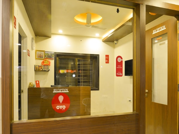 A reception area with a reception desk, a window with blinds, a ceiling fan, and a wall-mounted television. - Hotel Midtown Inn (Trimbak)
