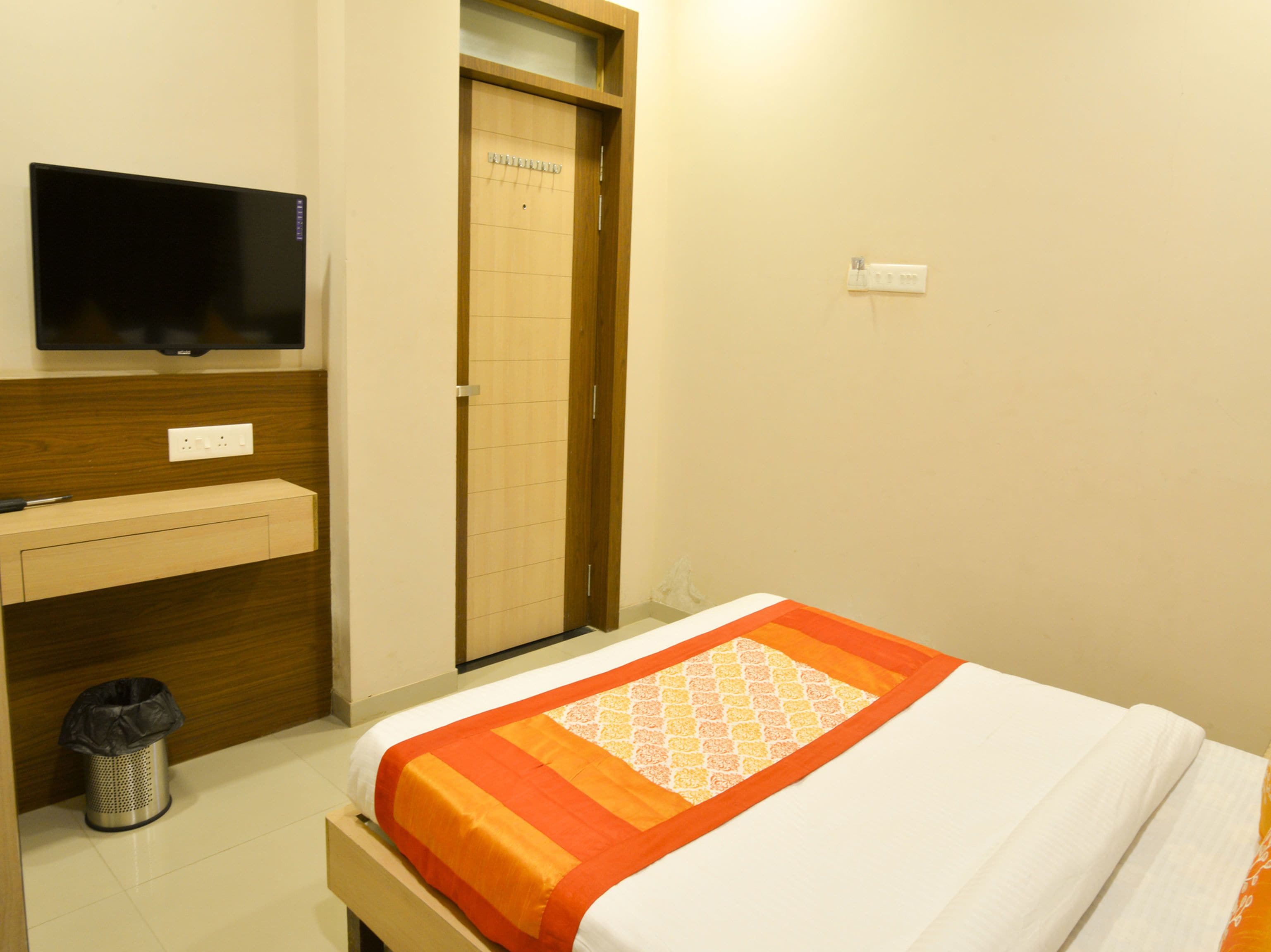 A hotel room with a bed, a TV mounted on the wall, a wooden cabinet, a trash bin, and a door with a sign.