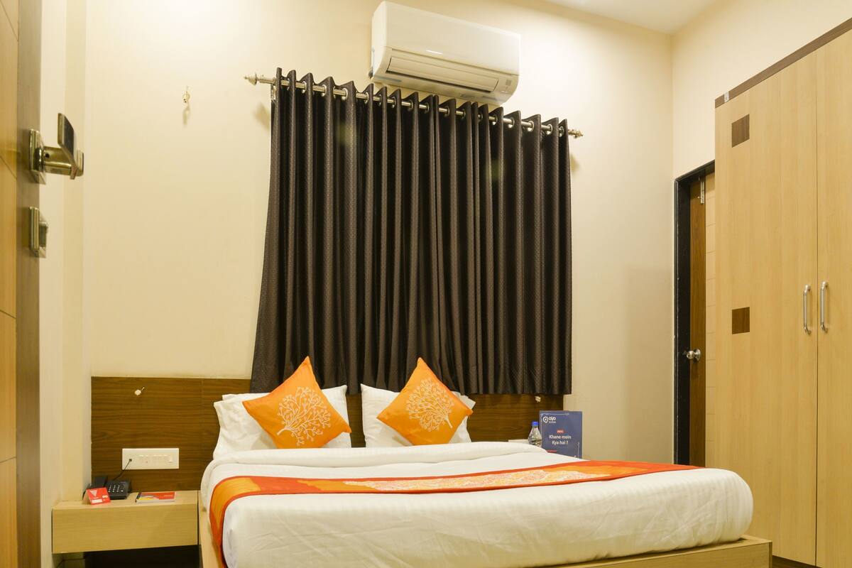 A hotel room with a bed, an air conditioning unit, a nightstand with a phone and a water bottle, and a wardrobe.