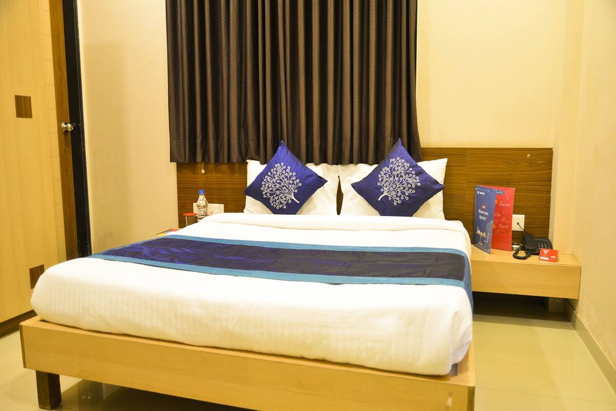 A hotel room with a wooden bed, dark curtains, a bedside table with a phone and a bottle, and a nightstand with a red box.