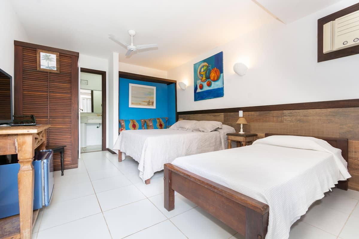 Une chambre d’hôtel avec deux lits, un bureau, une chaise, un ventilateur de plafond et des œuvres d’art accrochées aux murs.